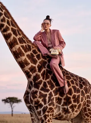 Model in a dusty pink suit and tinted sunglasses perched on a giraffe at dusk, holding a cream rotary phone