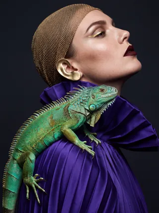 Model in profile wearing a gold mesh cap and purple pleated gown, a green iguana perched on her shoulder