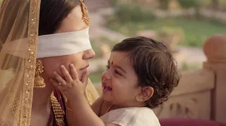Blindfolded woman in gold jewellery and a cream sari cradling a laughing toddler who touches her cheek, sunlit garden