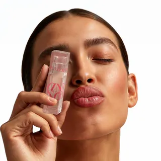 Woman with slicked-back hair winks and pouts, holding a clear pink-labelled lip gloss tube against her cheek