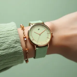Wrist wearing a rose-gold watch with mint leather strap and a gold chain bracelet against a sage green backdrop