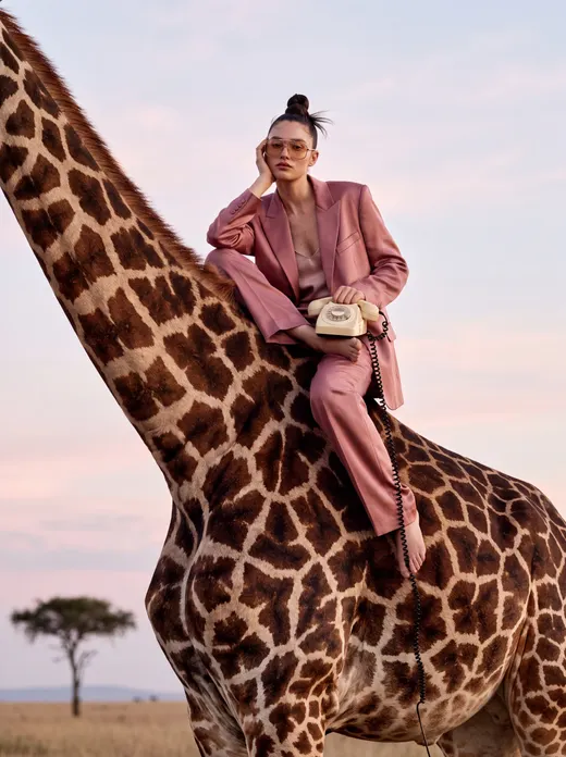 Model in a dusty pink suit and tinted sunglasses perched on a giraffe at dusk, holding a cream rotary phone