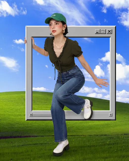 Woman in a green cap, olive top and wide-leg jeans stepping out of a retro desktop window over a grassy hill