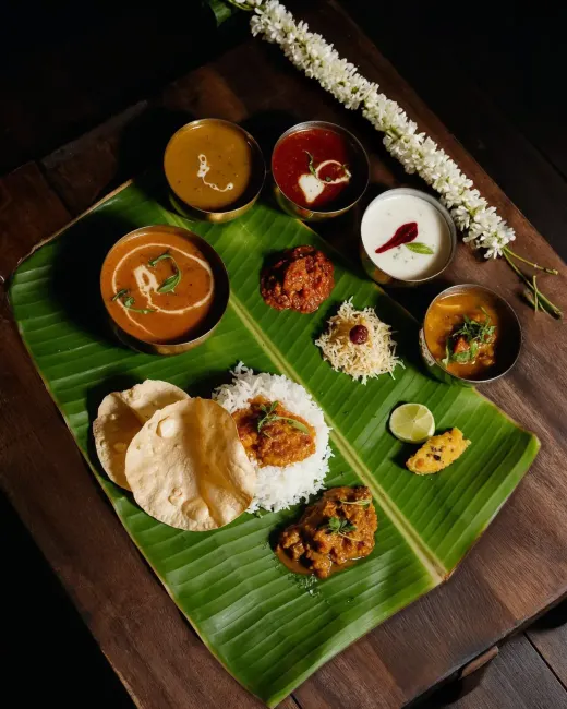 South Indian thali on a banana leaf — curries in brass bowls, rice, papadums and a jasmine garland on dark wood