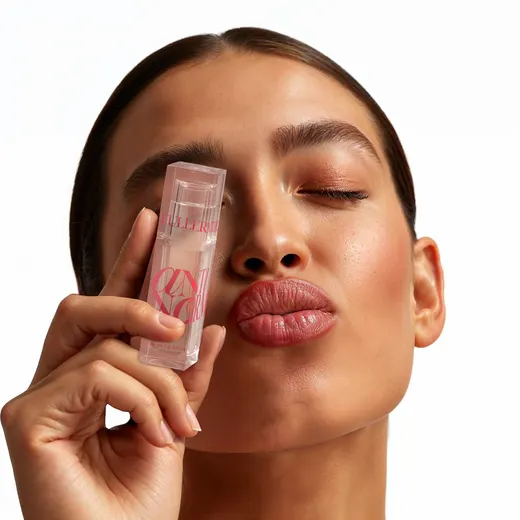 Woman with slicked-back hair winks and pouts, holding a clear pink-labelled lip gloss tube against her cheek
