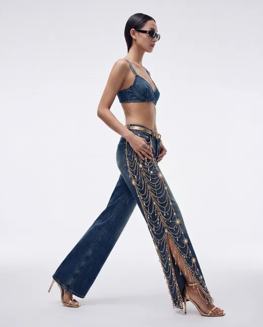 Model in a denim bralette and wide-leg jeans draped in gold chains, walking in profile against a pale studio backdrop